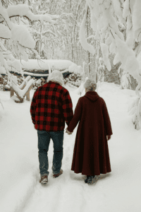 An elderly couple, Santa and Mrs. Claus, walk hand-in-hand through a snow-covered forest trail. Santa wears a red and black plaid flannel jacket, and Mrs. Claus wears a long burgundy wool cloak.