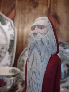 Close-up of a hand-carved wooden Santa figure with a long white beard and red coat, crafted by Santa Claus in a folk-art style and placed in front of vintage Christmas dishes.